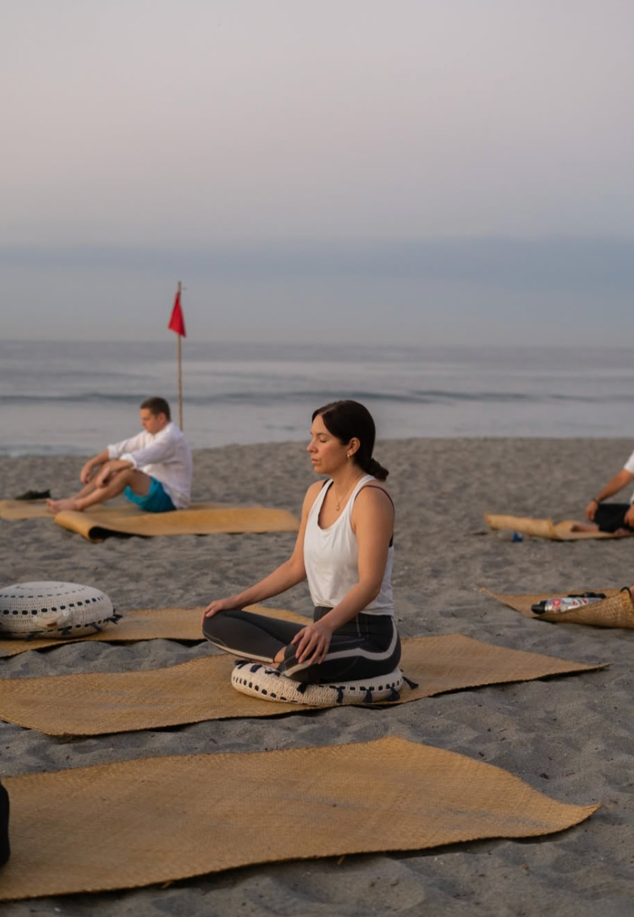 Personas sentadas con las piernas cruzadas sobre esterillas en la playa durante una sesión de yoga al amanecer en cangrejera cardedeu