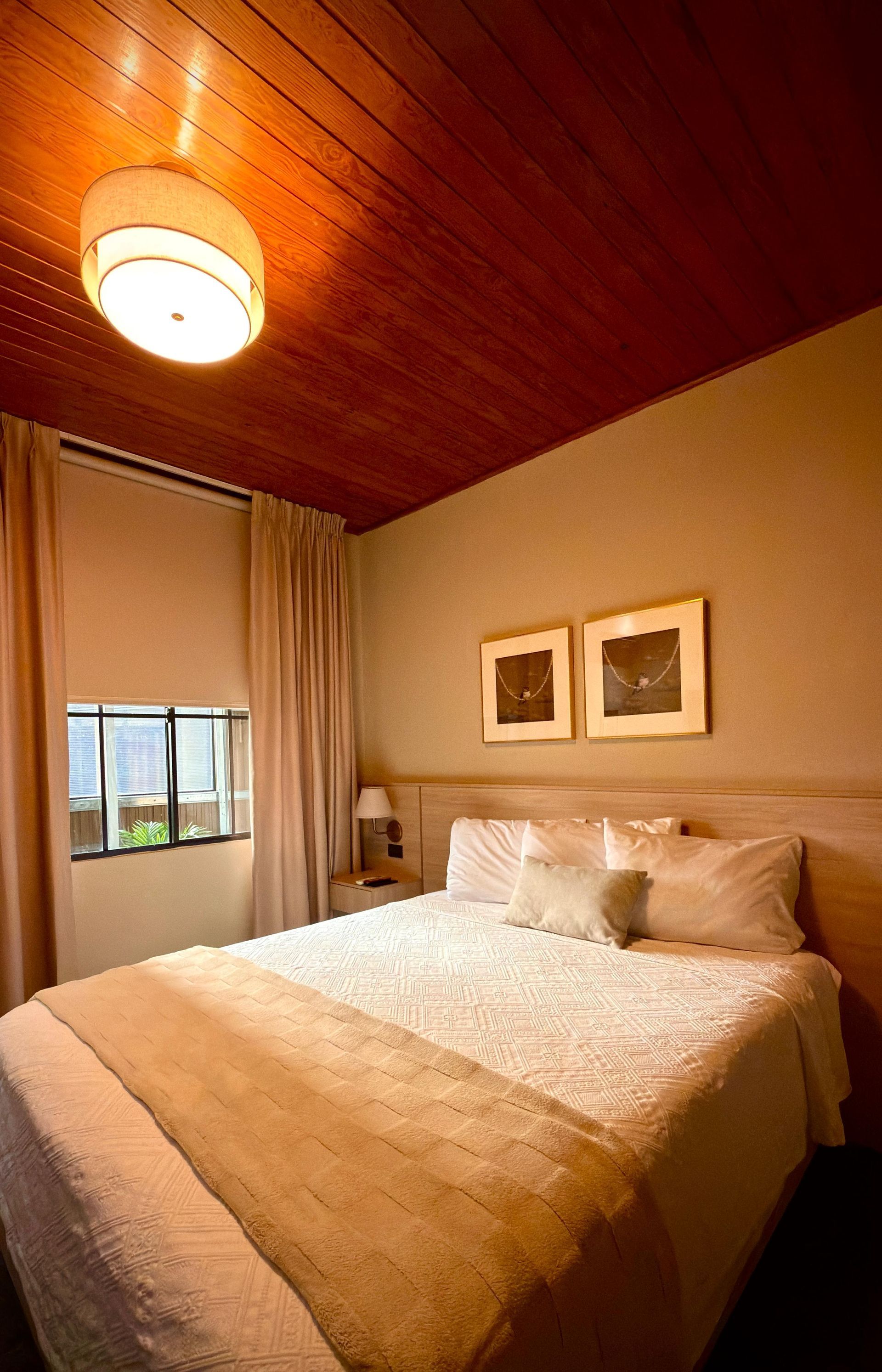 Habitación acogedora con cama, techo de madera cálida, paredes beige y ventana con cortinas en hotel cardedeu suites