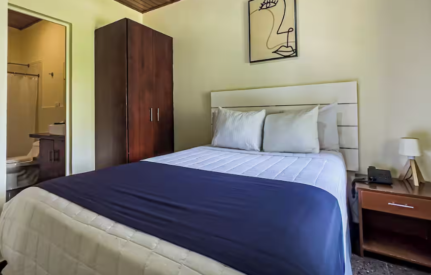 Habitación luminosa con una cama bien hecha, una manta azul oscuro, un armario de madera y una lámpara de mesilla de noche.