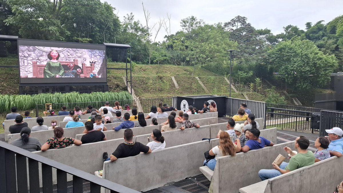 Personas sentadas al aire libre viendo una película en una pantalla grande en un entorno similar a un parque.