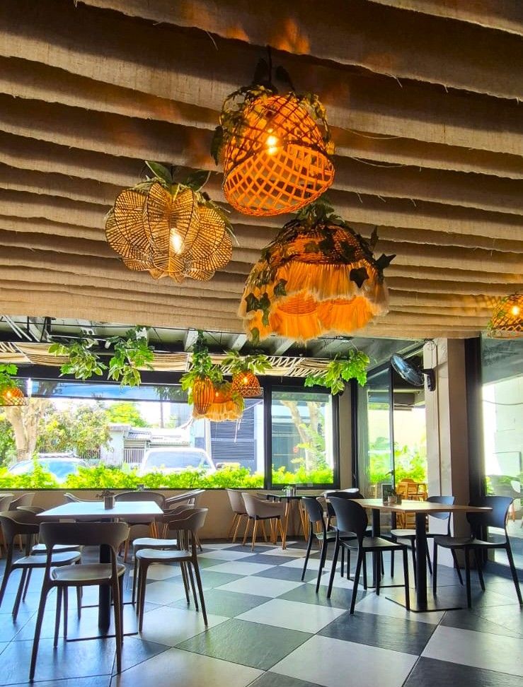 Cafetería elegante con lámparas colgantes de mimbre, suelo en blanco y negro y grandes ventanales con vistas a zonas verdes.