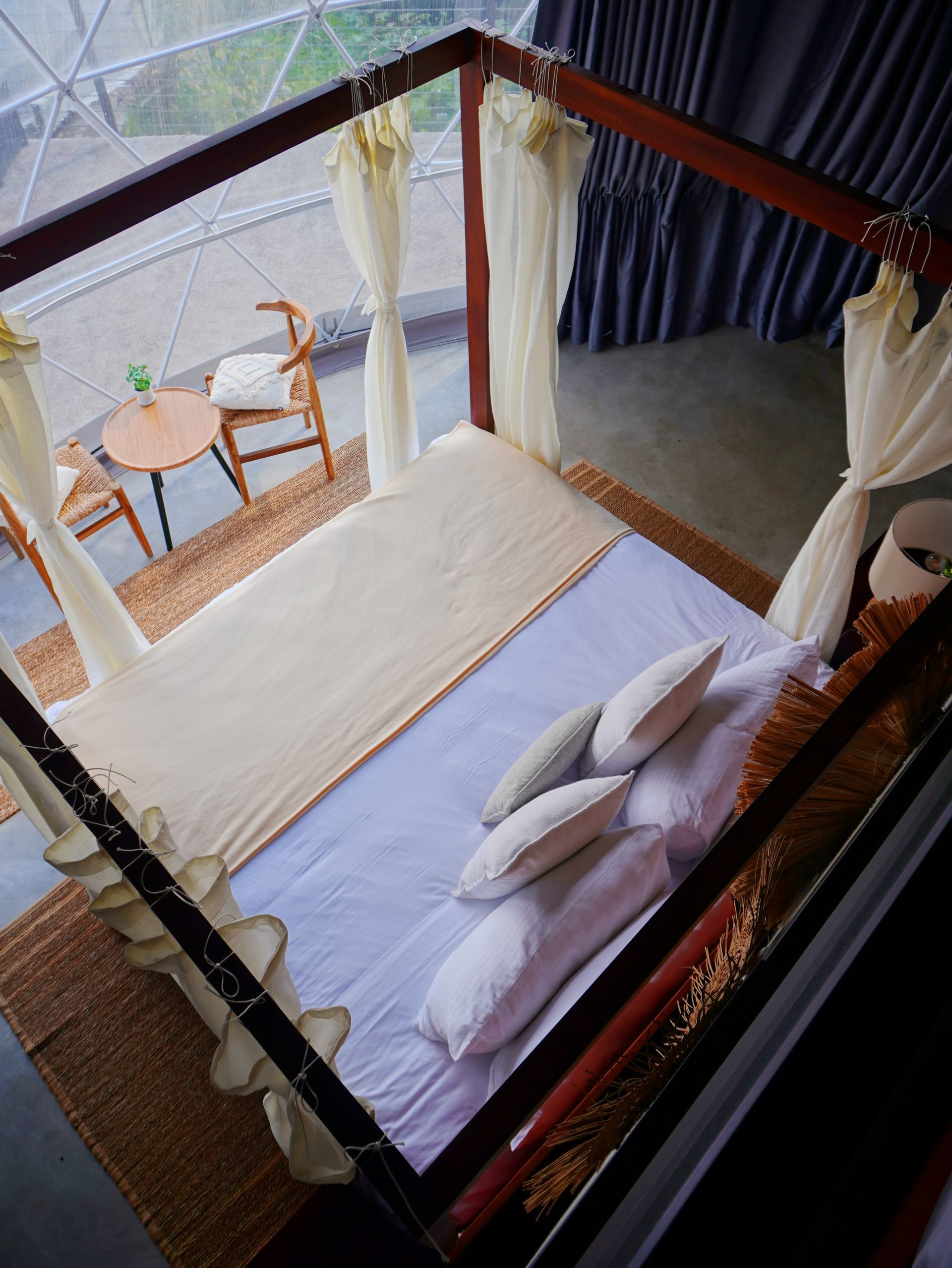 Cama con dosel, sábanas blancas y almohadas junto a un pequeño balcón con taburetes y cortinas.