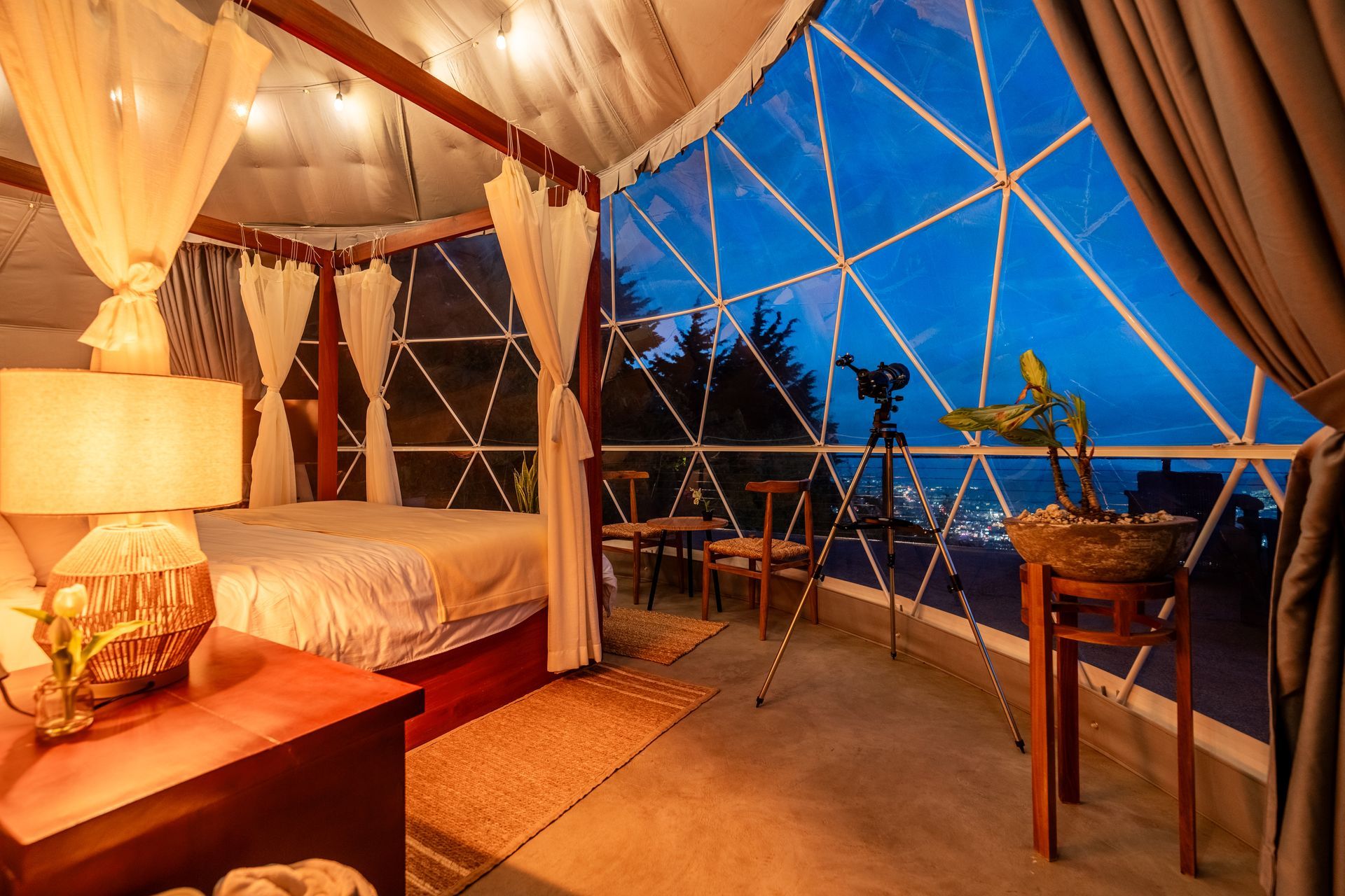 Acogedora habitación tipo tienda de campaña con cama king size, lámpara cálida y vistas azules al atardecer a través de paredes transparentes.