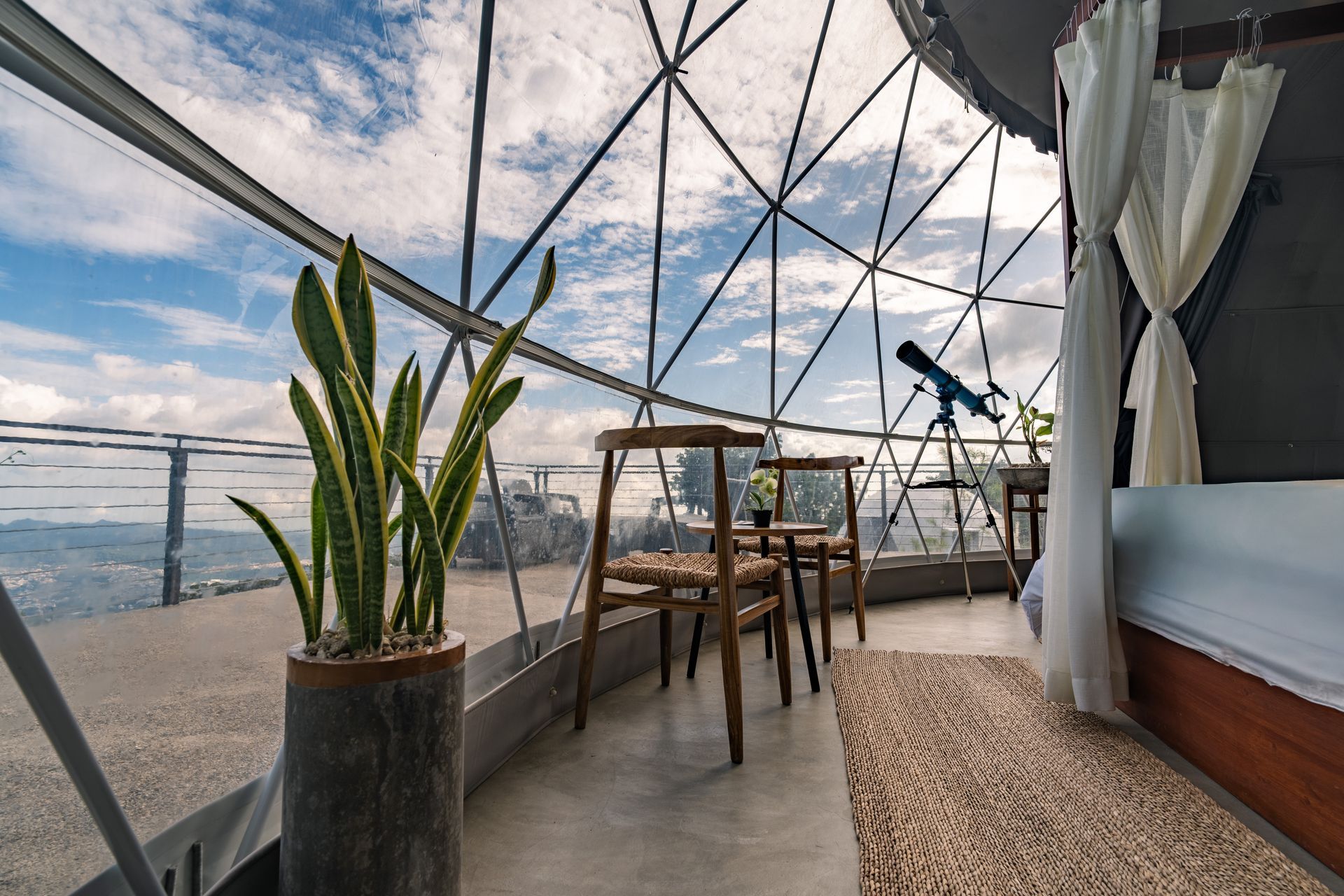 Salón al aire libre con planta en maceta, mesa de madera, sillas y vista al océano bajo una cúpula de cristal.