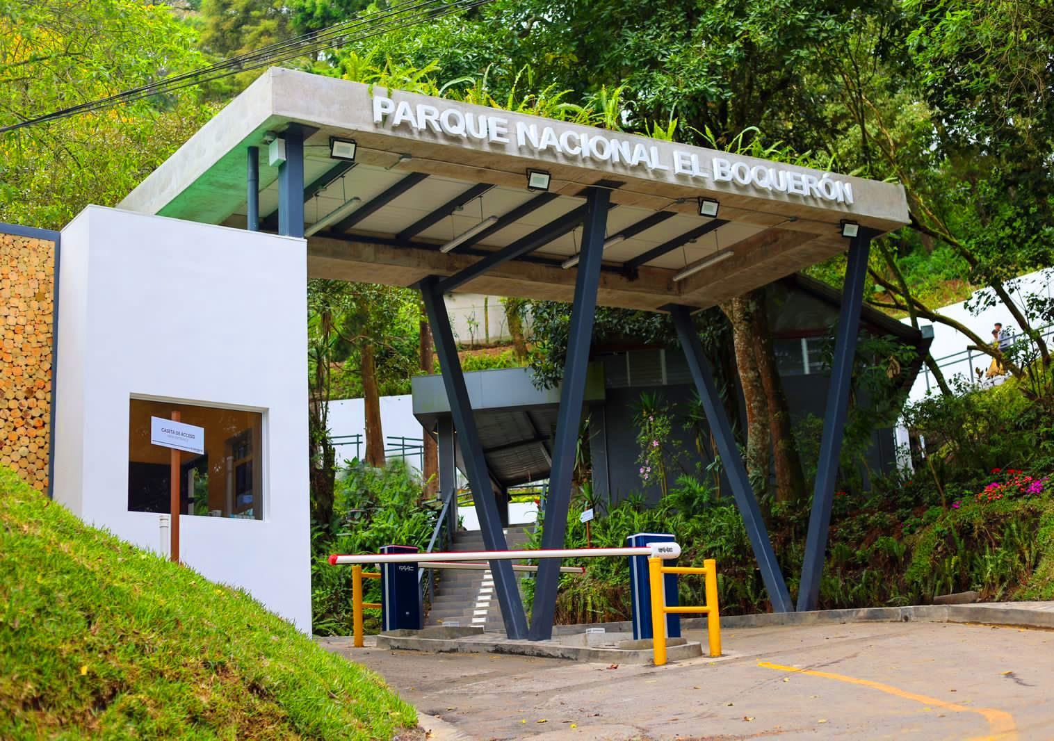 Entrada al parque con un letrero blanco, techo inclinado y árboles que rodean la puerta.
