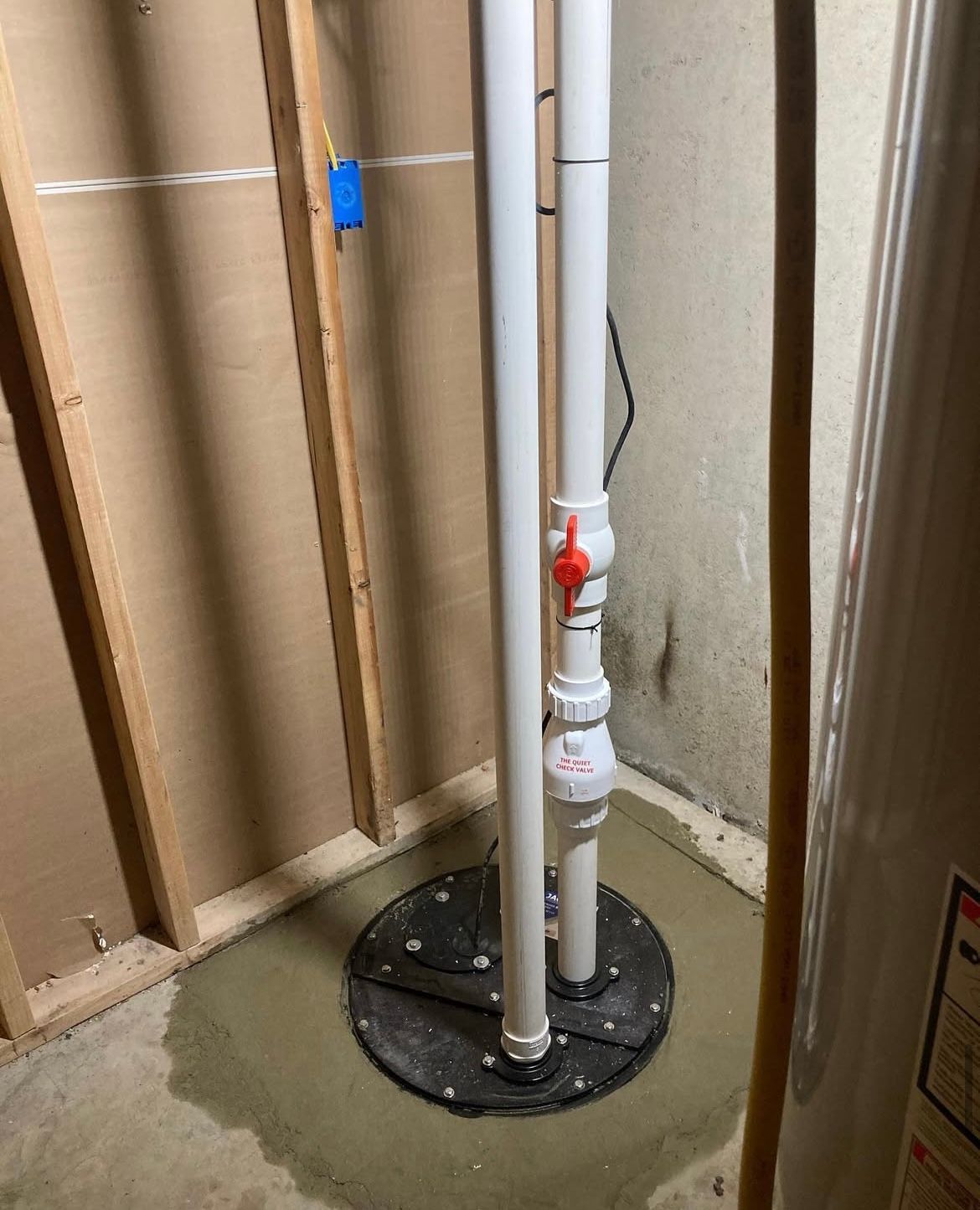 Sump pump with white pipes, a red valve, and black base in a concrete-floored basement setting.