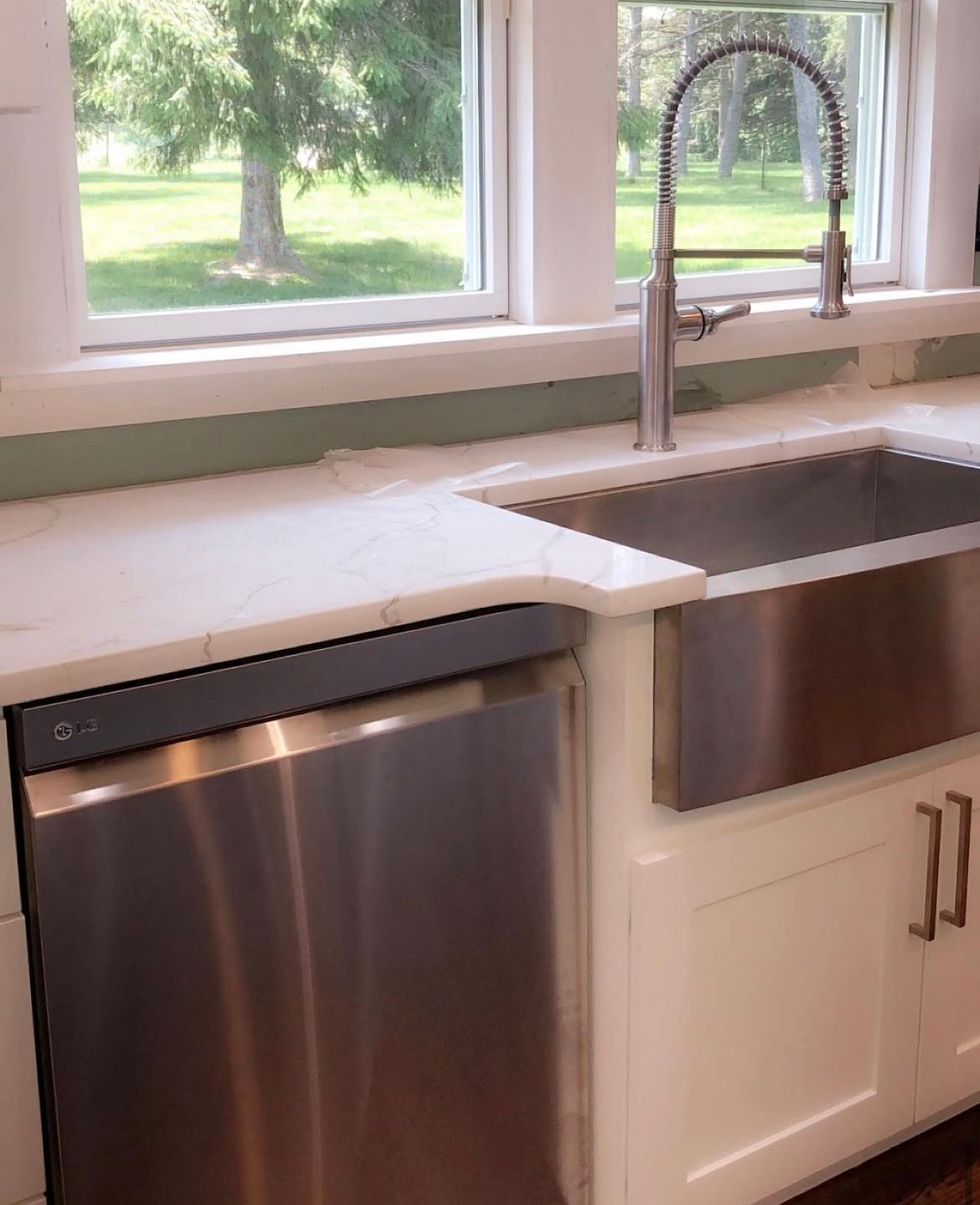 Stainless steel kitchen sink and dishwasher with white cabinets and a window overlooking a green lawn.