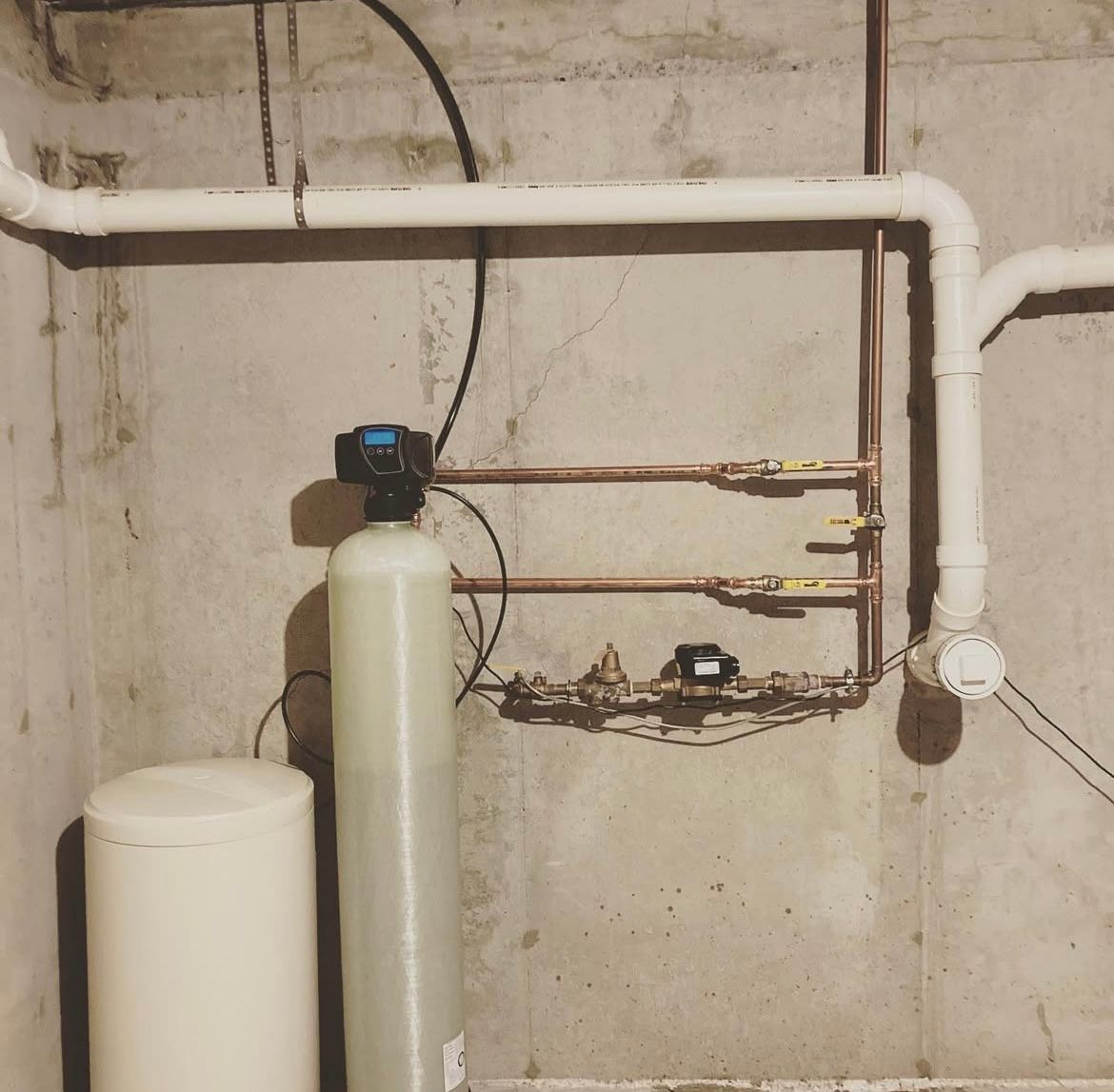 Water softener system with a white tank and copper piping, set against a concrete wall.