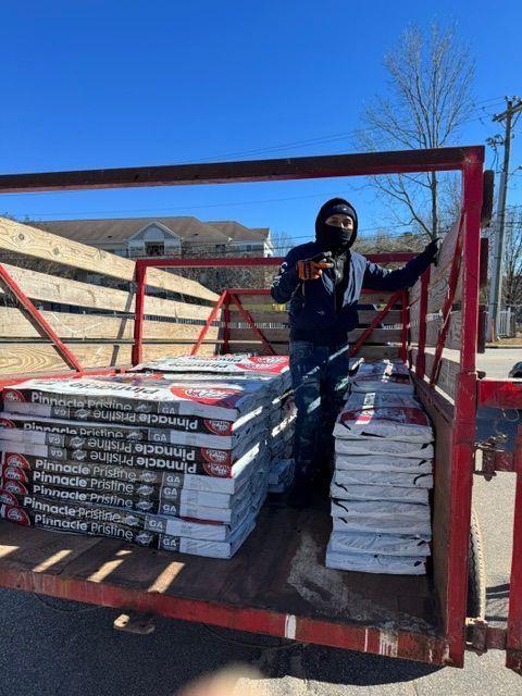 Worker standing on a red trailer loaded with stacked roofing shingles under a clear blue sky