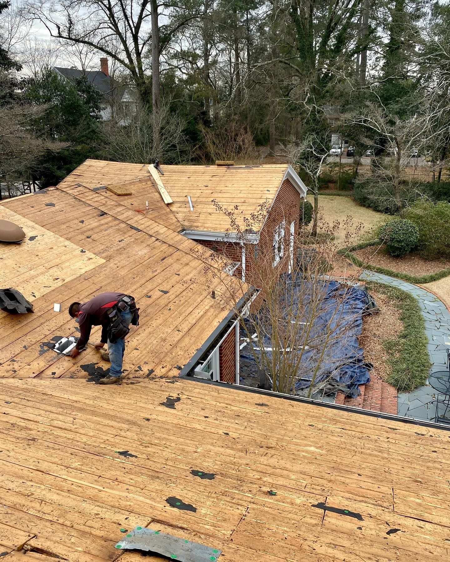 Roofers working on a tan shingle roof beside trees and a brick building
