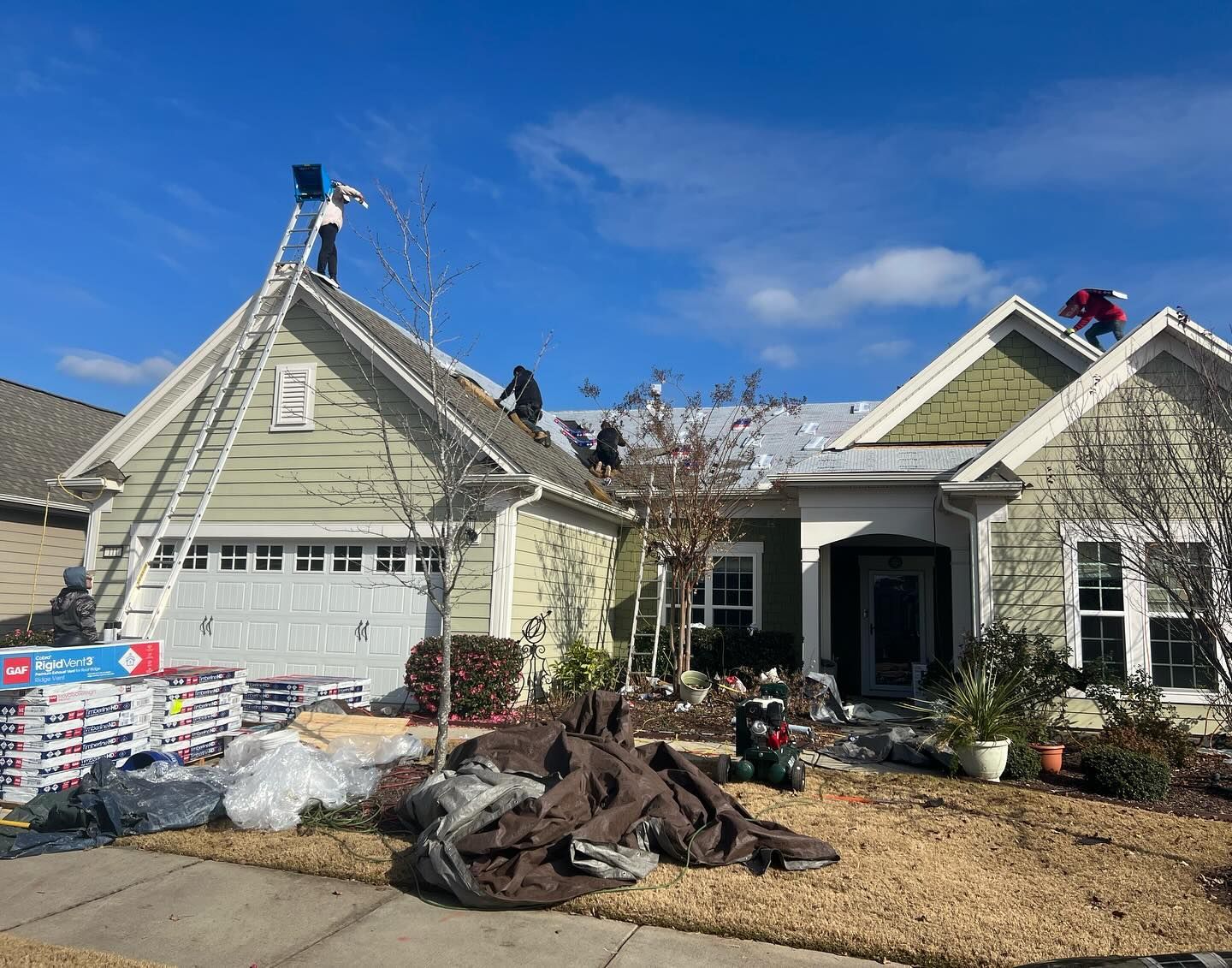 Workers on ladders repairing a house roof, with shingles and materials piled in the front yard.