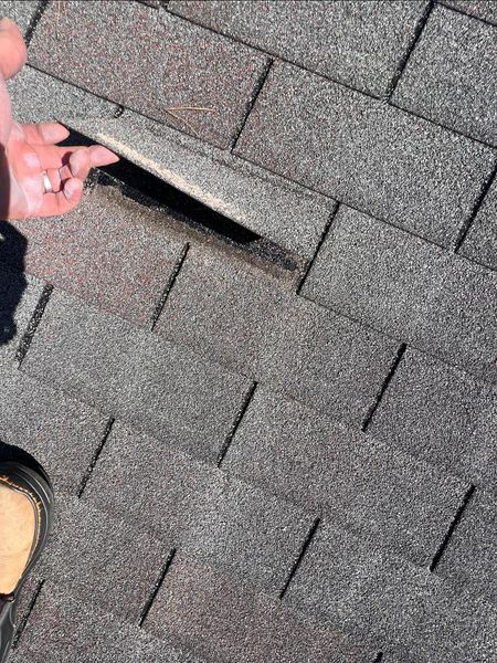Hand lifting a damaged asphalt shingle on a roof, revealing a gap underneath.