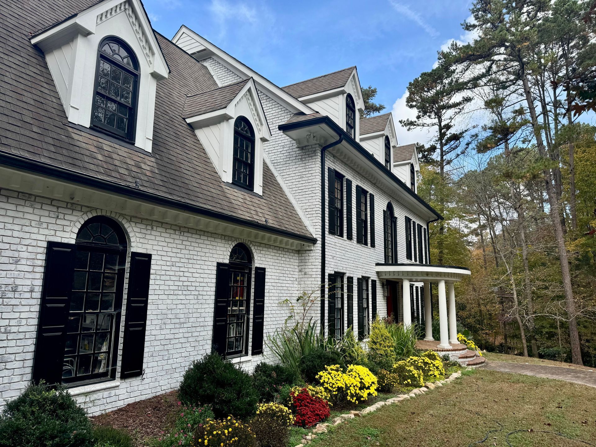 Large white brick house with black shutters, arched windows, and colorful shrubs along the front