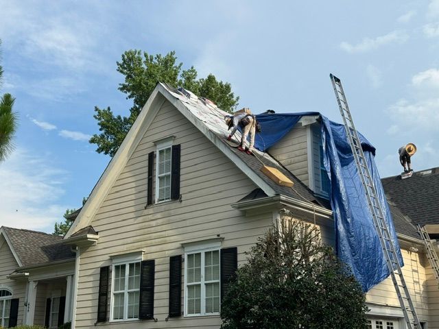 Roofers repairing blue-tarped house roof with ladders and shingles