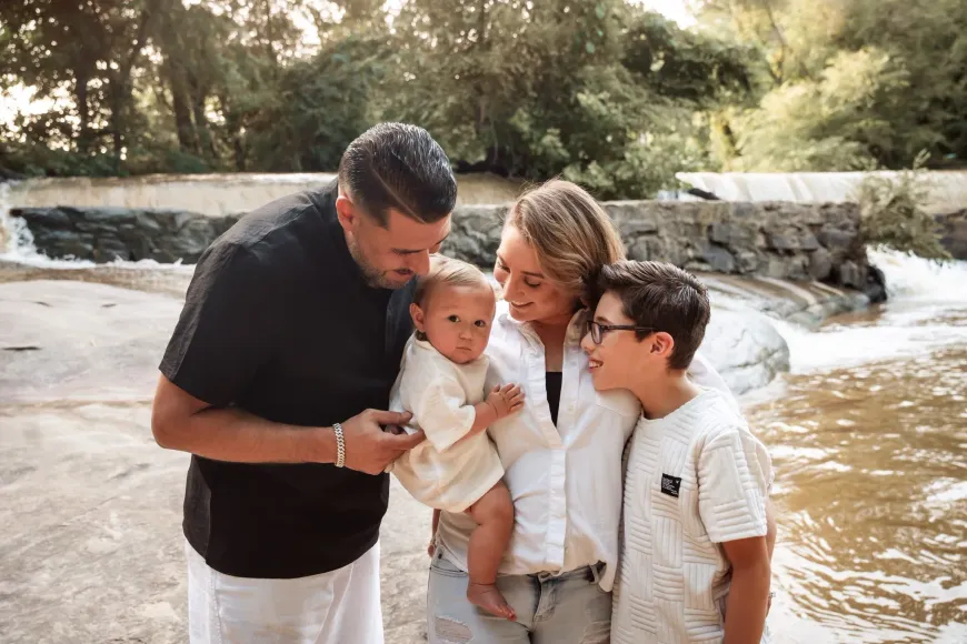Family standing by a waterfall, smiling and holding a baby outdoors.