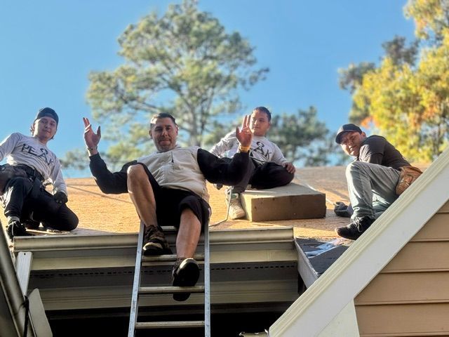 Four people sitting on a roof edge, waving down with ladders and a blue sky behind them.