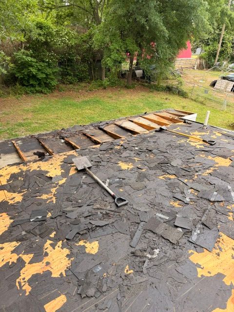 Damaged roof with torn black shingles, exposed wood, and a rake on top near trees and grass.