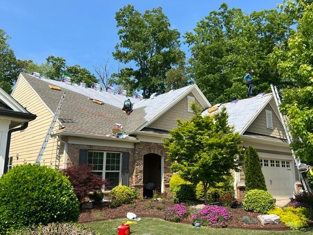 Suburban house with workers on the roof, beige siding, white garage, and a landscaped front yard.