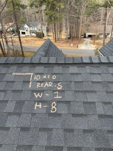 Roof shingles marked with chalk measurements near a roof ridge, overlooking a wooded residential yard.