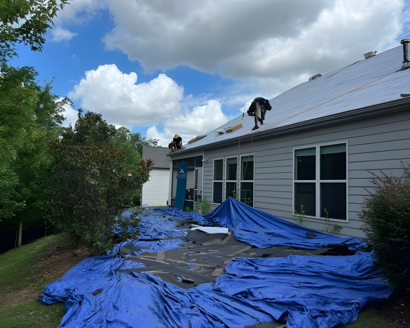 Workers on a tarp-covered roof repairing shingles on a house under a cloudy sky