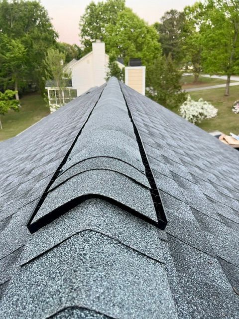 Rooftop ridge vent on a gray shingled roof with trees and a house in the background