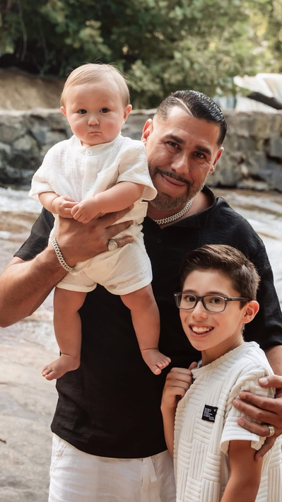 A man holds a baby while a smiling child stands beside them outdoors.