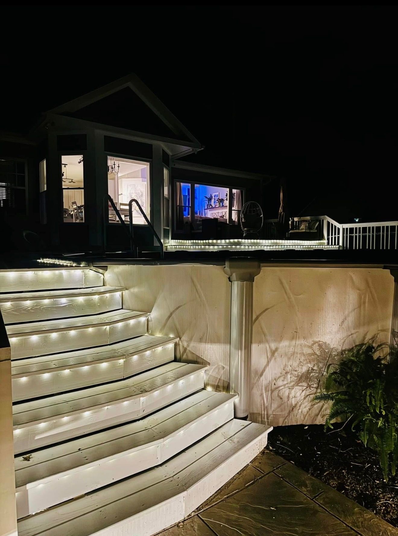 Lit white outdoor steps leading to a house deck at night, with lit windows and a railing.