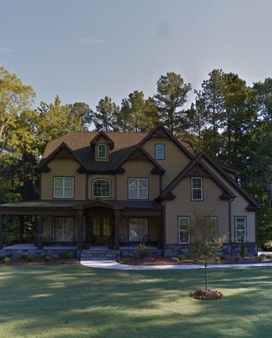 Large beige suburban house with a front porch, green lawn, and tall trees in the background.