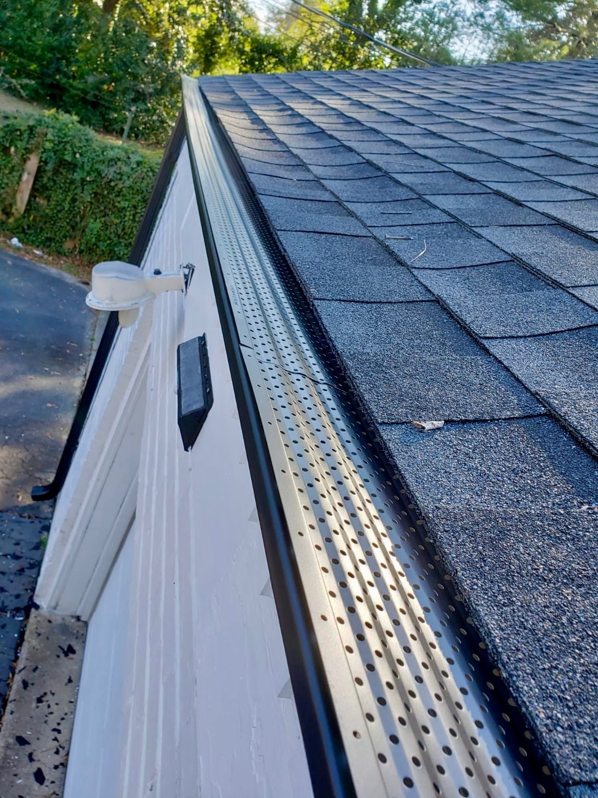 Close-up of a house roof with dark shingles, white siding, and a gutter along the edge.