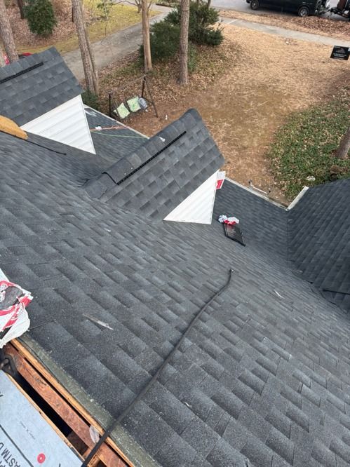 Workers repairing a gray shingled roof beside a wooded yard