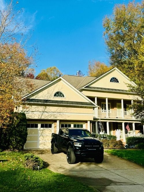 Black SUV in driveway of a beige two-story house with a porch and autumn trees under a blue sky