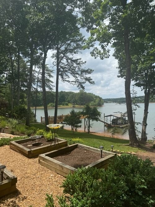 Wooded lakeside view with garden beds, gravel path, and docks under cloudy sky
