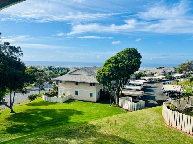 Sunny suburban hillside with a white house, green lawn, trees, and ocean view under a blue sky