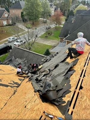 Worker repairing damaged roof with exposed plywood and missing shingles in a residential neighborhood