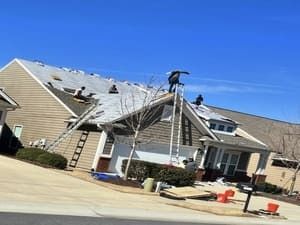 Roofers working on a beige house roof with ladders and tarps in a sunny suburban driveway