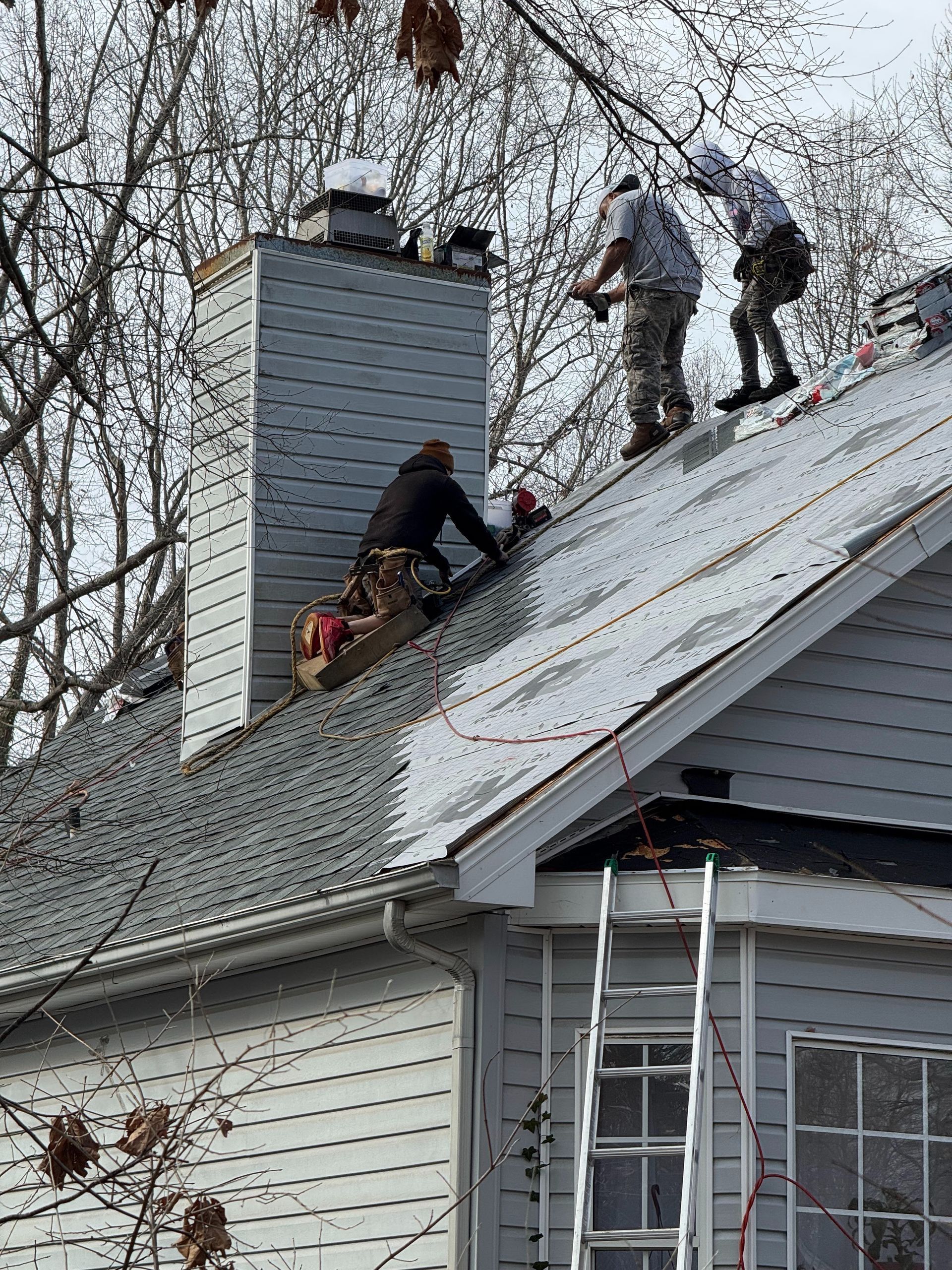 Workers repairing a gray house roof near a chimney, with a ladder leaning below.
