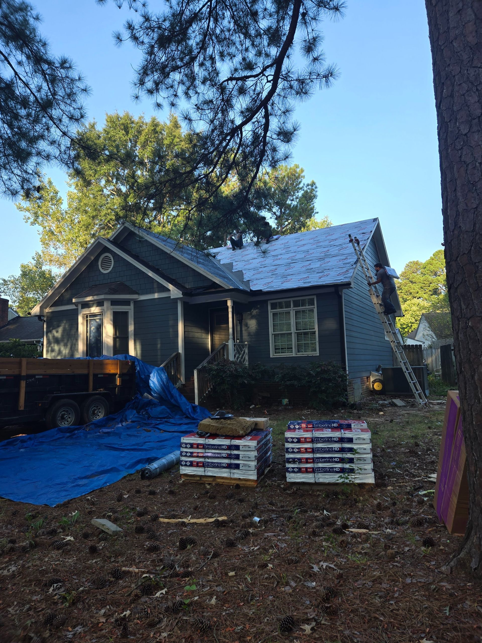 House with roof being repaired, blue tarp over a truck, and stacked materials in front yard.
