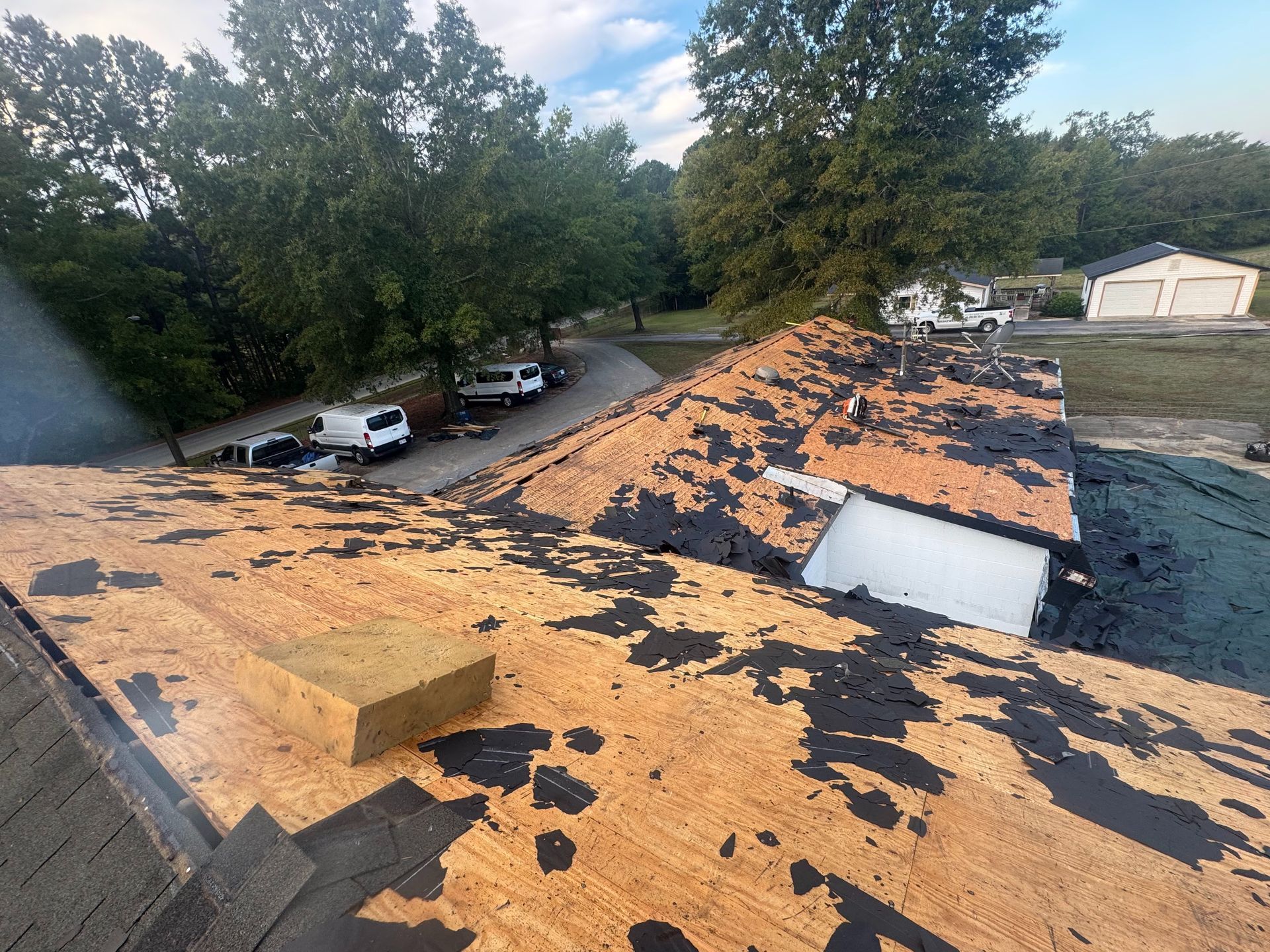 Damaged roof with missing shingles and debris, overlooking trees and parked vehicles below.