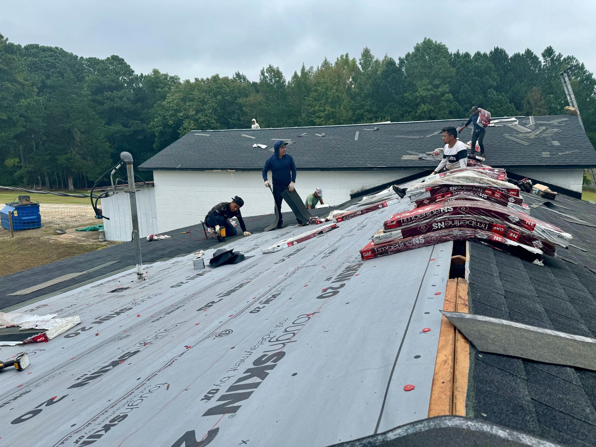 Workers installing roofing on a damaged house with exposed sheathing and shingles, seen from the roofline.
