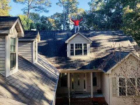 Person in red shirt standing on a house roof among dormers and trees