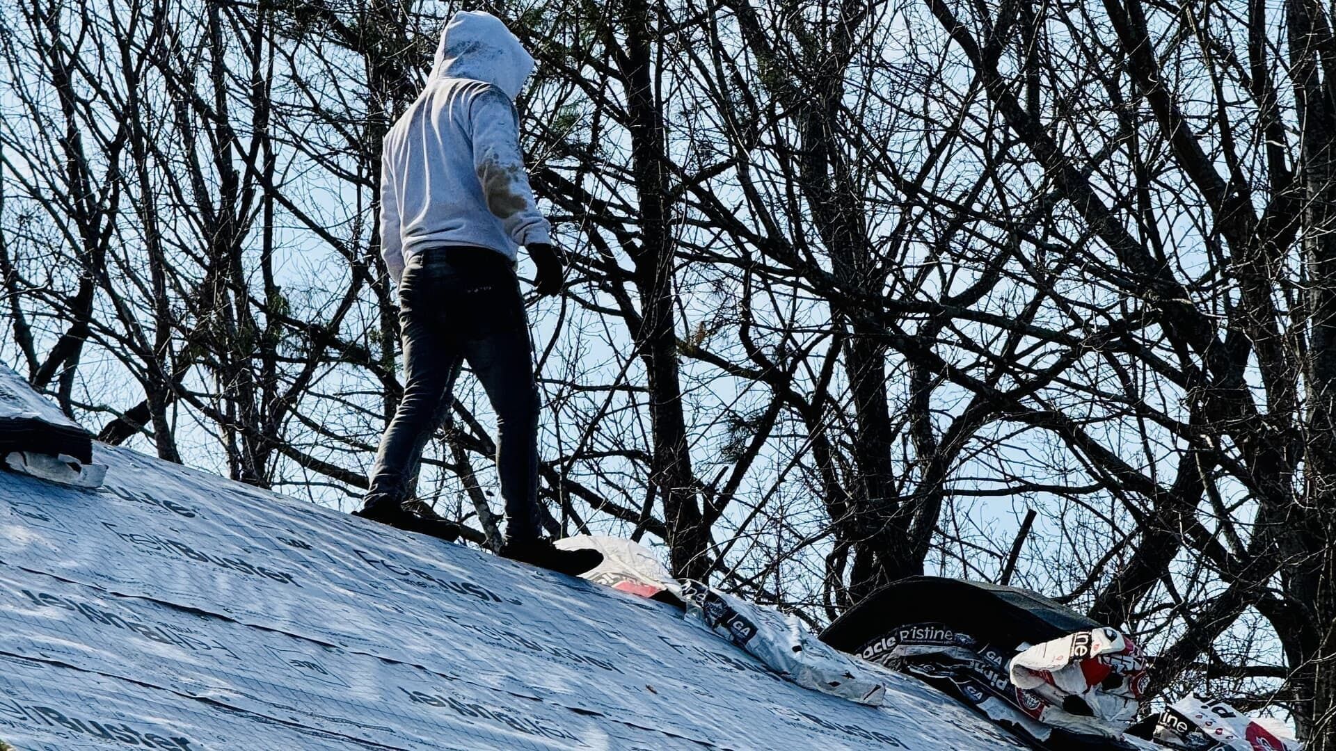 Person in a white hoodie stands on a snowy roof among bare trees.