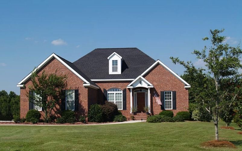 Brick house with a dark roof and white trim on a green lawn under a clear blue sky.