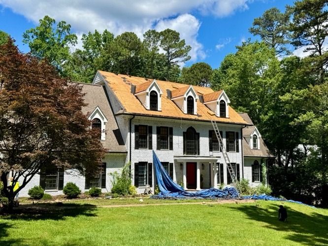 Large white house with orange roof and blue tarp in front, surrounded by trees and a green lawn