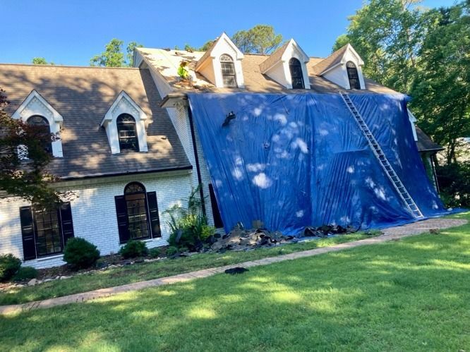 House with blue tarp covering roof and upper wall, surrounded by green lawn and trees