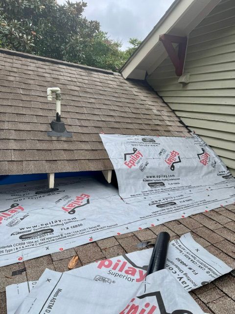 Roof under repair with silver underlayment exposed and brown shingles above a wall and vent pipe