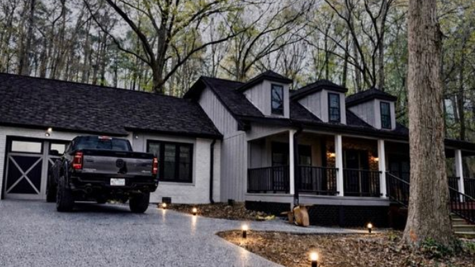 White house with black roof and dormers, front porch, driveway, and parked black pickup in a wooded yard.
