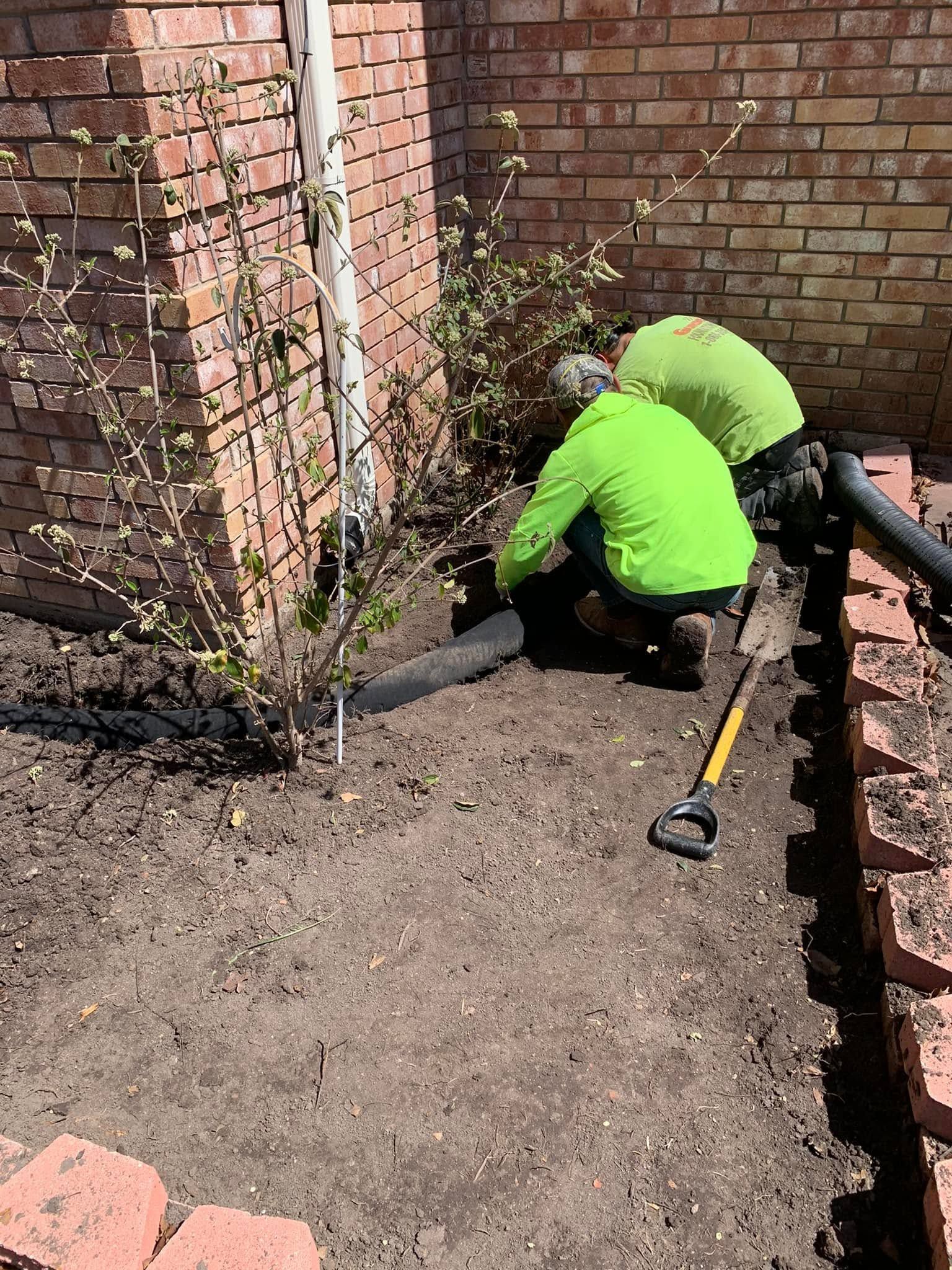 Two workers in bright green shirts kneel in a brick courtyard, digging near a wall with a shovel.