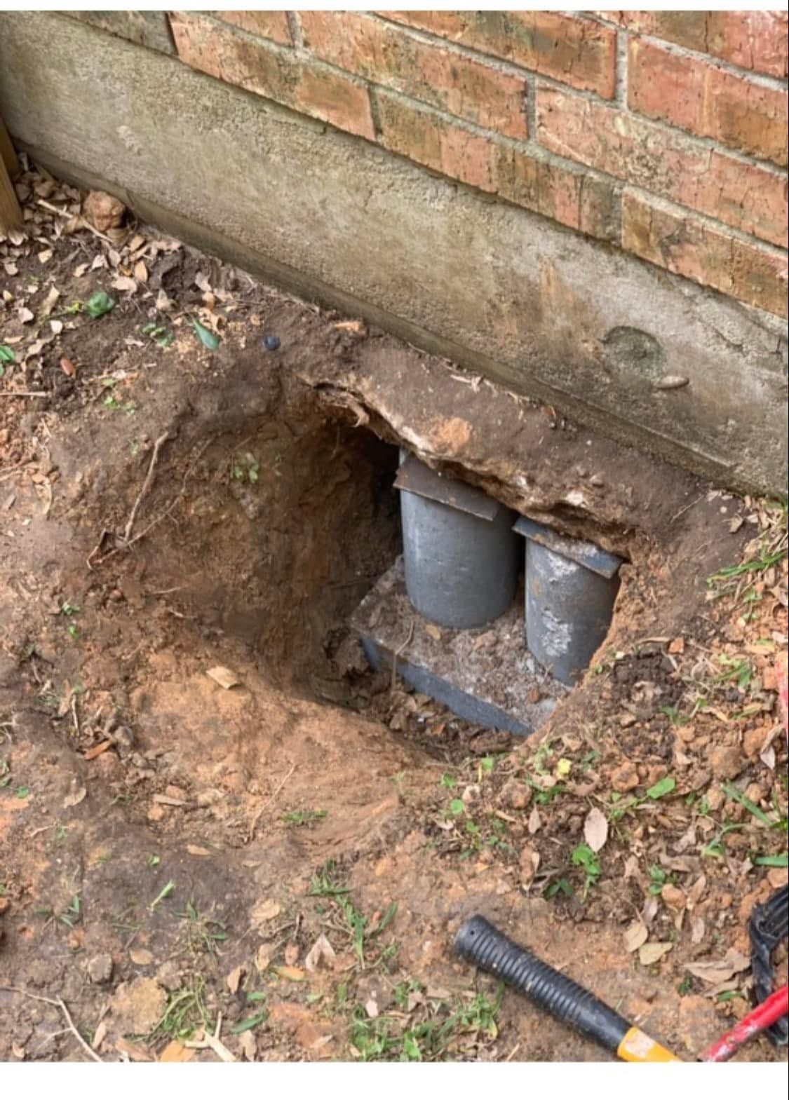 Exposed utility pipes in a dug-out hole beneath a brick wall outside a house