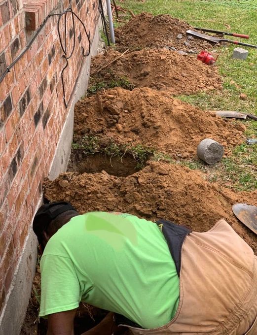 Worker digging a trench beside a brick wall, with tools and piles of dirt on grass.