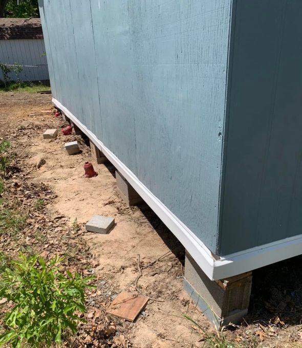 Blue-sided shed on cinder blocks beside a dirt strip and scattered bricks along a wall.